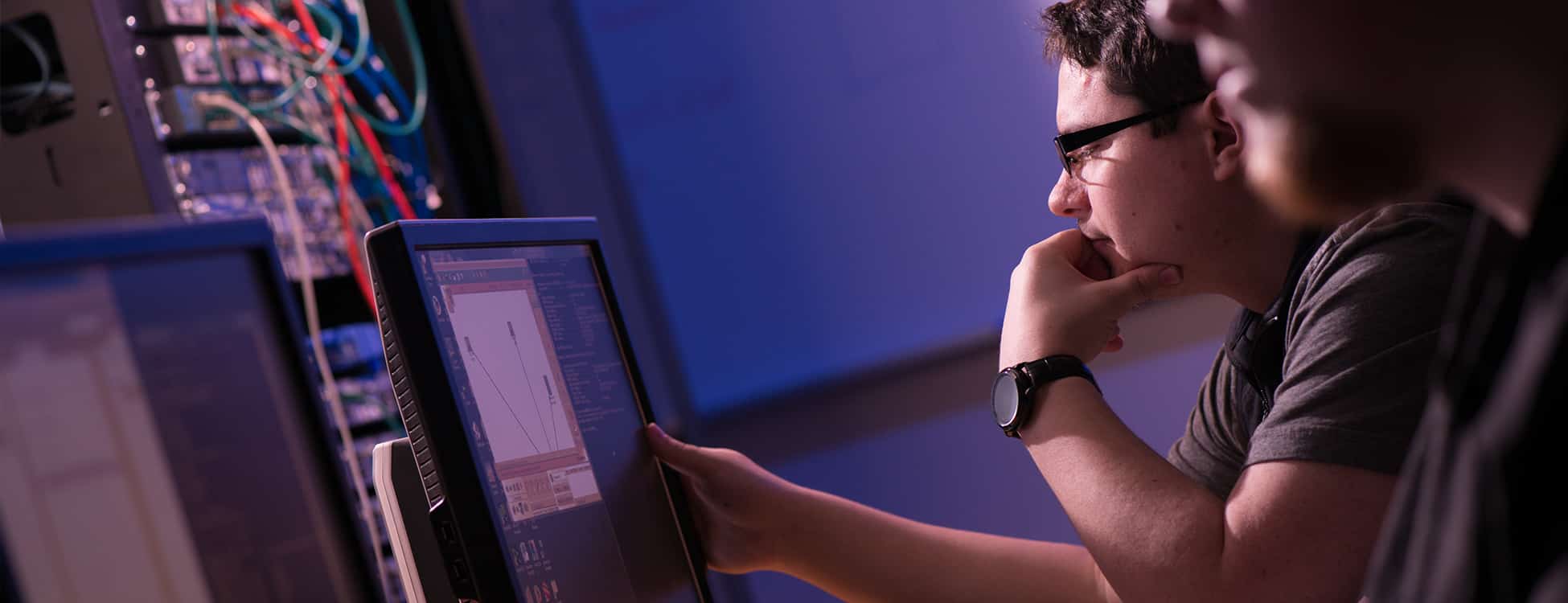 Male student works on computer screen in dimly lit room with colorful wires