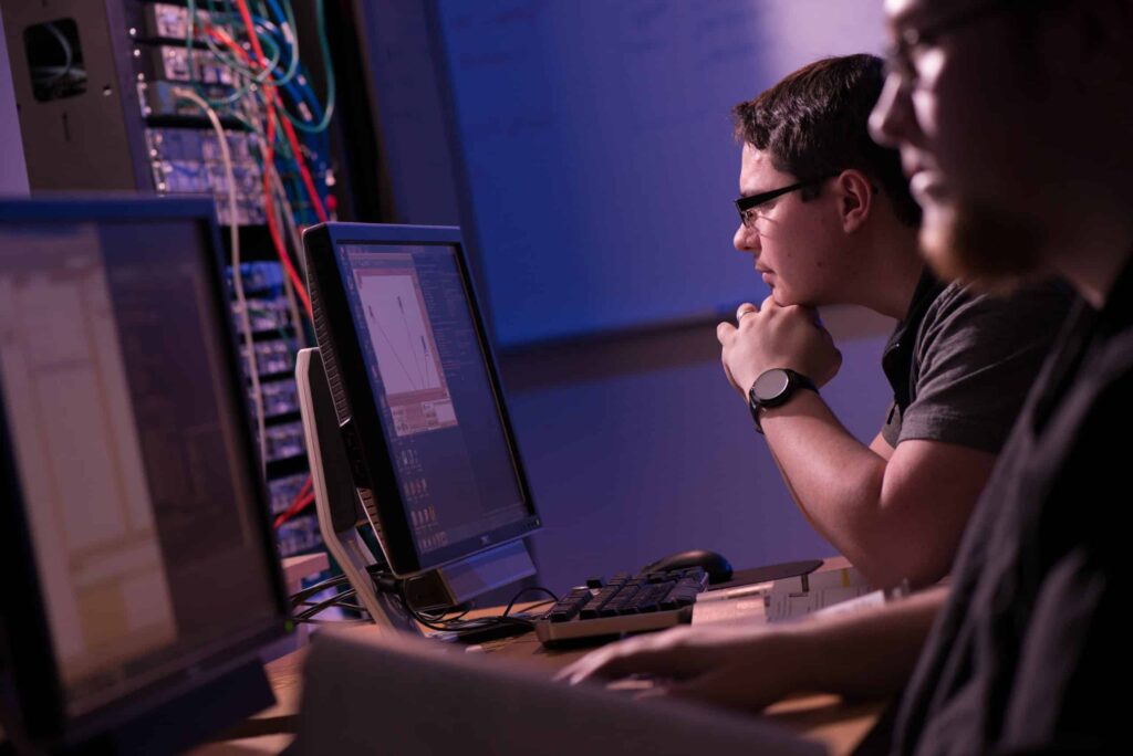 Two male students work on computer screen coding in a dimly sit room with computer wires and books.