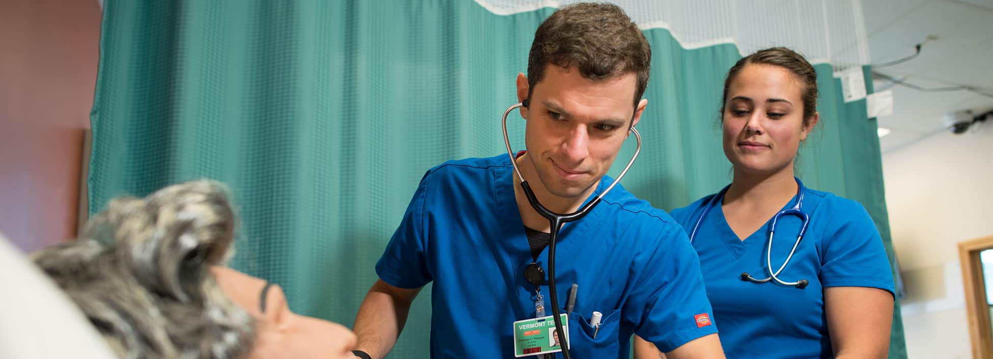 A male nursing student examines a simulation patient while a female nursing student looks on, Williston campus