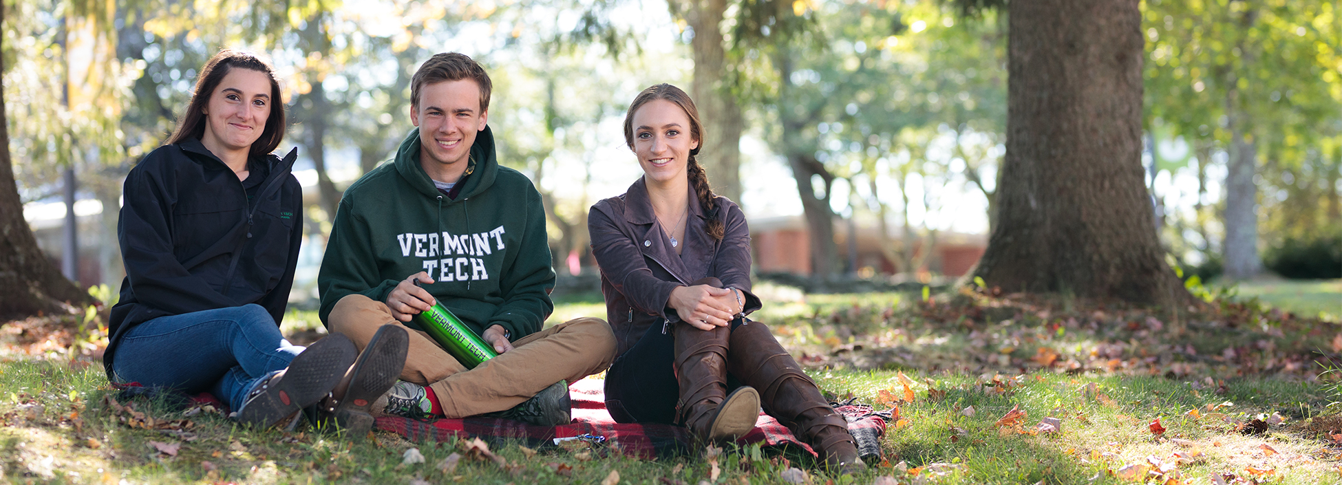 three students enjoy an afternoon on the lawn under the trees, smiling, Randolph Center campus