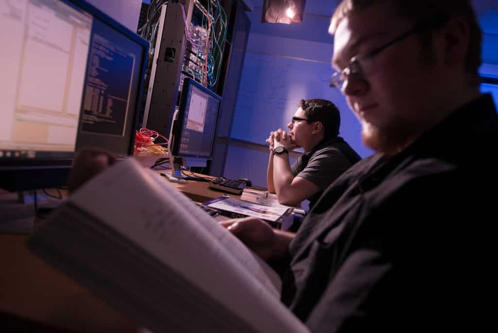 Two male students work on computer screens in a dark room, coding, computer wires, looking at screens