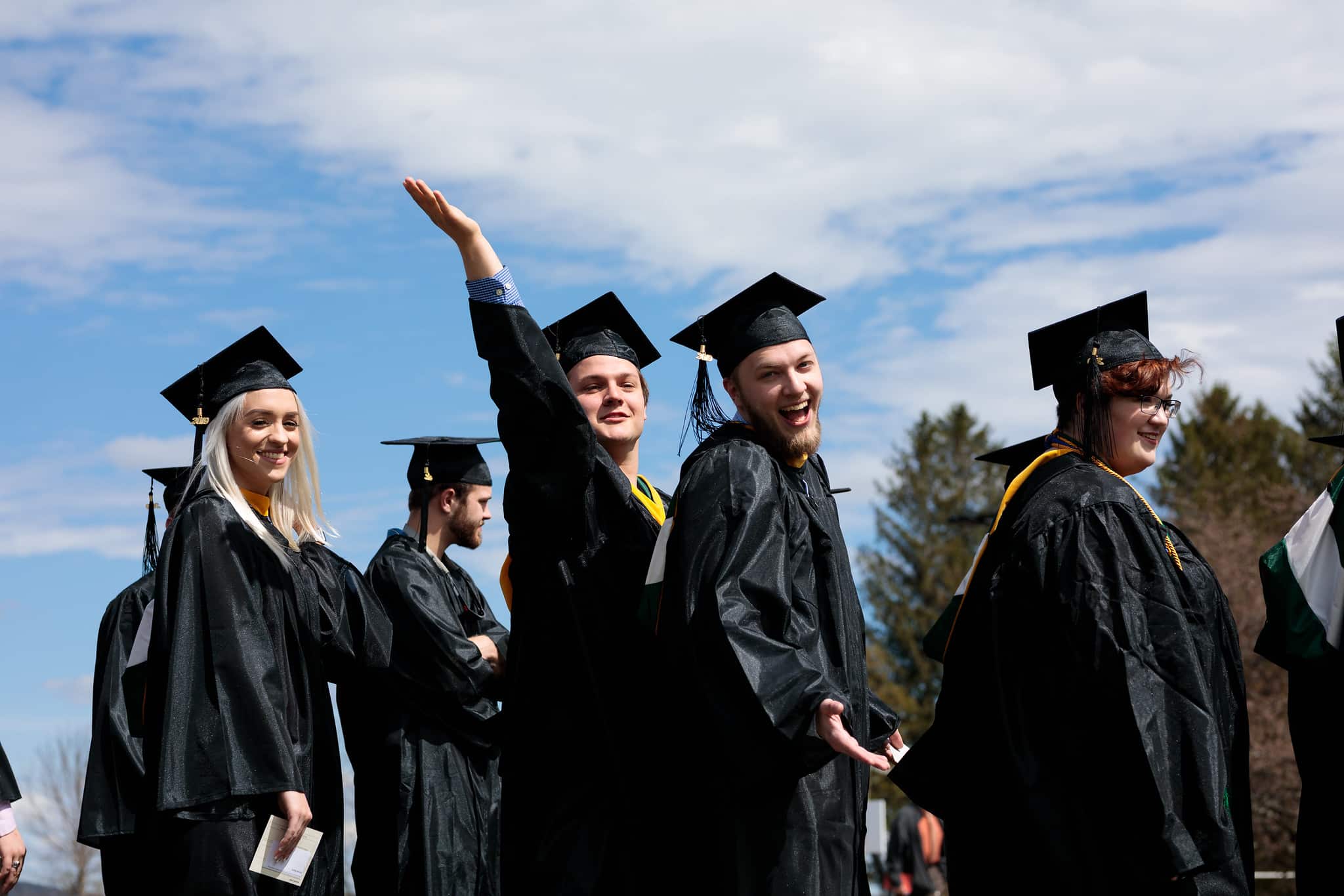 A recent Vermont Tech grad is waving as he processes with his fellow graduates in the graduation ceremony, wearing regalia